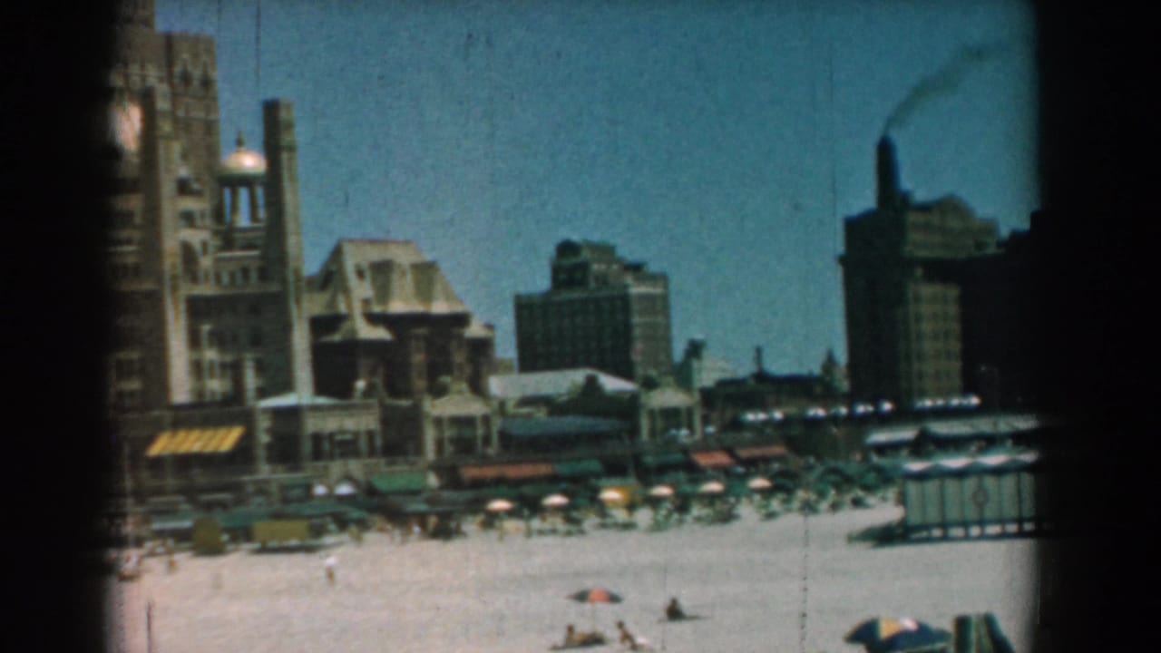 Sandy beach with distant people, umbrellas, large waterfront buildings, and a smoking stack.