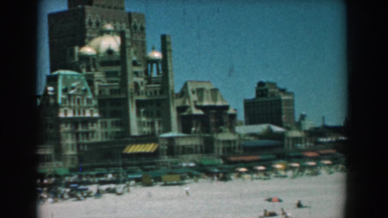 Wide beach scene with ornate buildings, umbrellas, cars, and smoke rising behind the skyline.