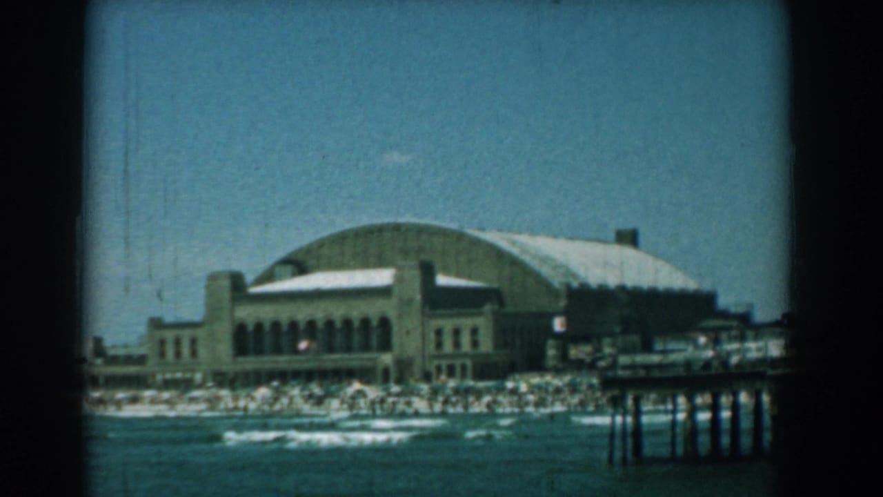 Distant waterfront building with arched roof behind a crowded beach and low waves.