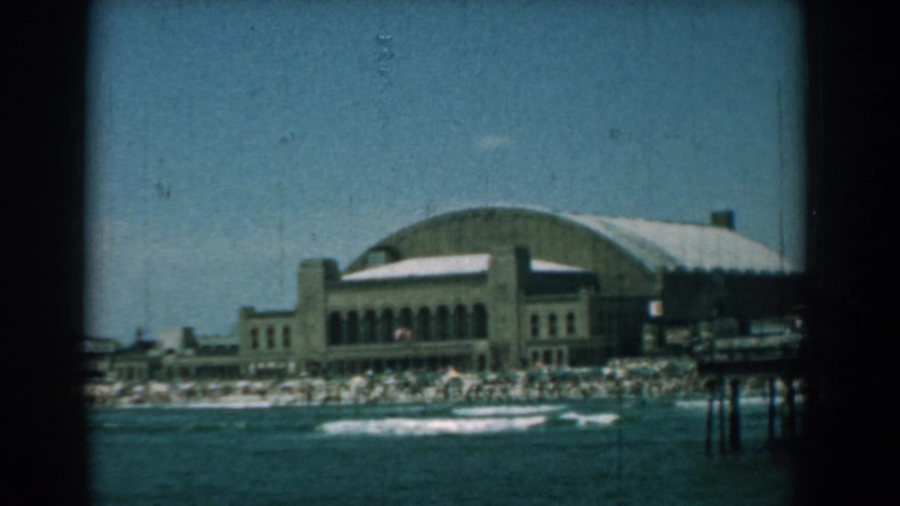 Large arched-roof building along the shore with beach crowds and waves in front.