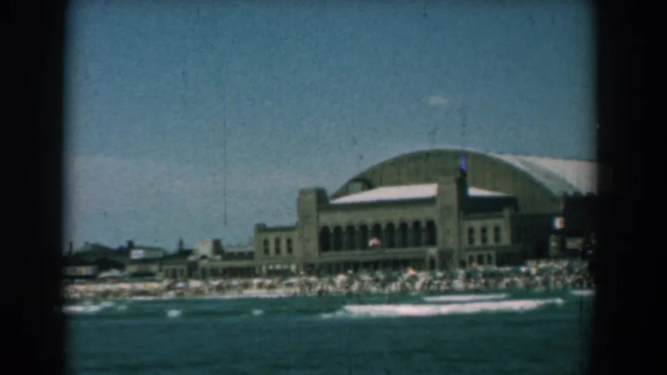 1958 home-movie frame of the Yorktown shoreline, with a large beachside building, crowded sand, rolling surf and soft 8mm color.