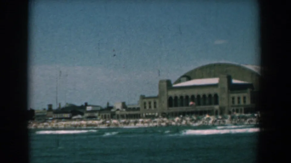 Wide 1958 Yorktown shoreline view with the arched-roof waterfront building partly to the right, crowds on the beach and surf in the foreground.