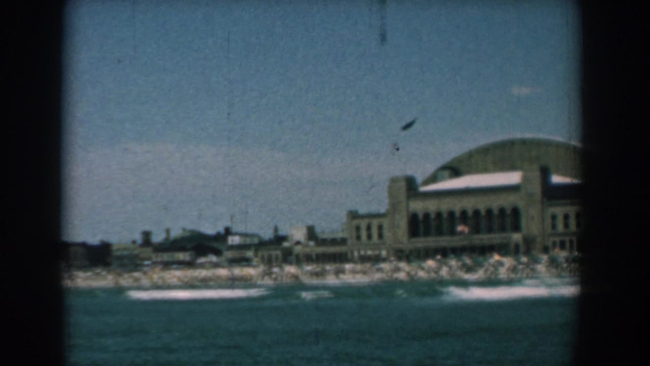 Shoreline resort building behind surf and beachgoers across blue water.