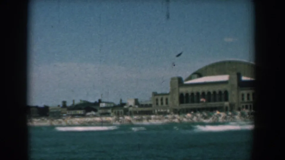 1958 Yorktown, Virginia home-movie frame showing blue water, breaking waves, a crowded beach, and a large curved-roof waterfront building.