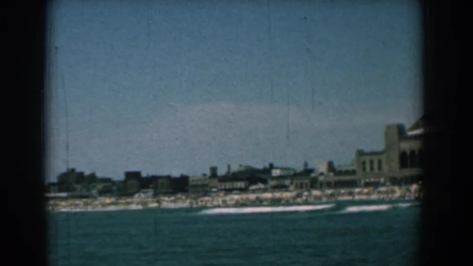 Distant 1958 home-movie view of the Yorktown shoreline, with turquoise water, white surf, beachgoers, and the resort building partly cropped at right.