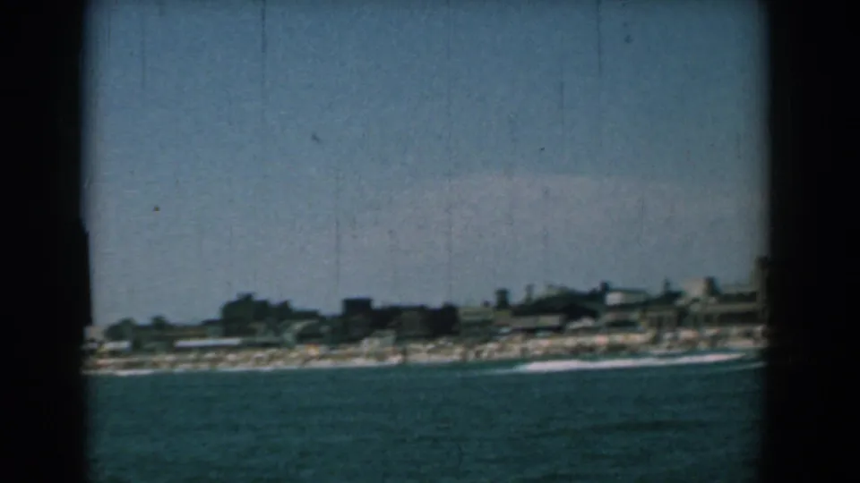 Final 1958 Yorktown waterfront frame with open blue water in the foreground, a narrow beach, low shore buildings, and small figures along the sand.