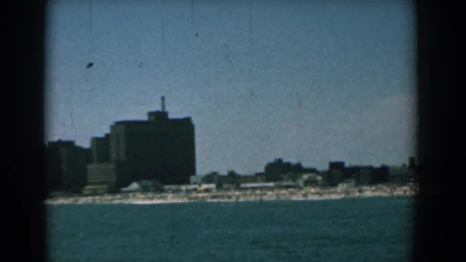 1958 coastal city view from offshore, showing a large dark building, neighboring structures, and a pale beach strip.
