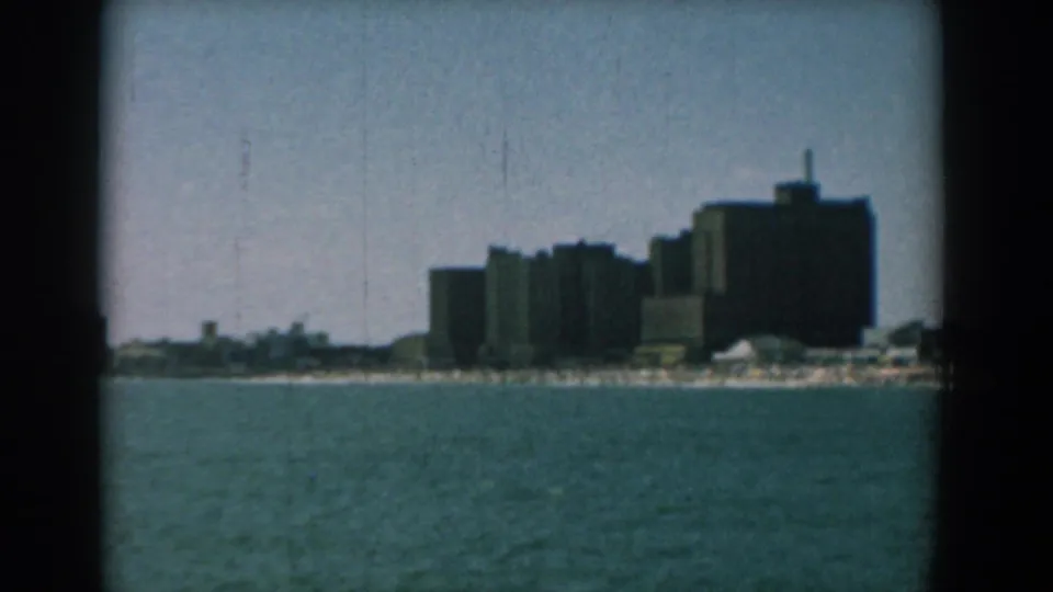 1958 home-movie still of a distant Yorktown building cluster along the shoreline, framed by 8mm gate edges.
