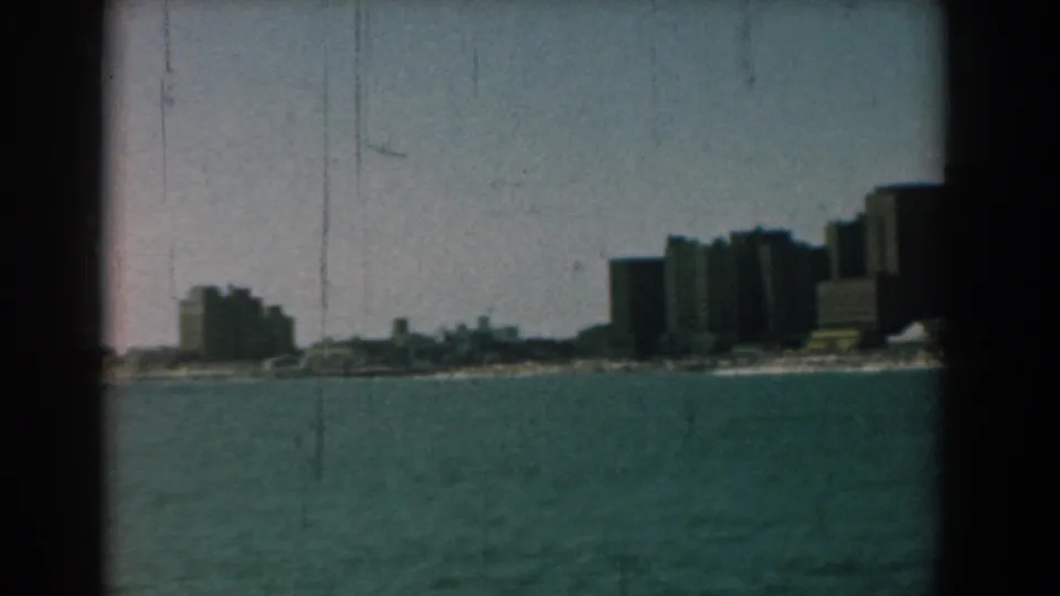 The frame holds on a distant coastal cityscape across blue-green water, with several mid-rise buildings along the shore.