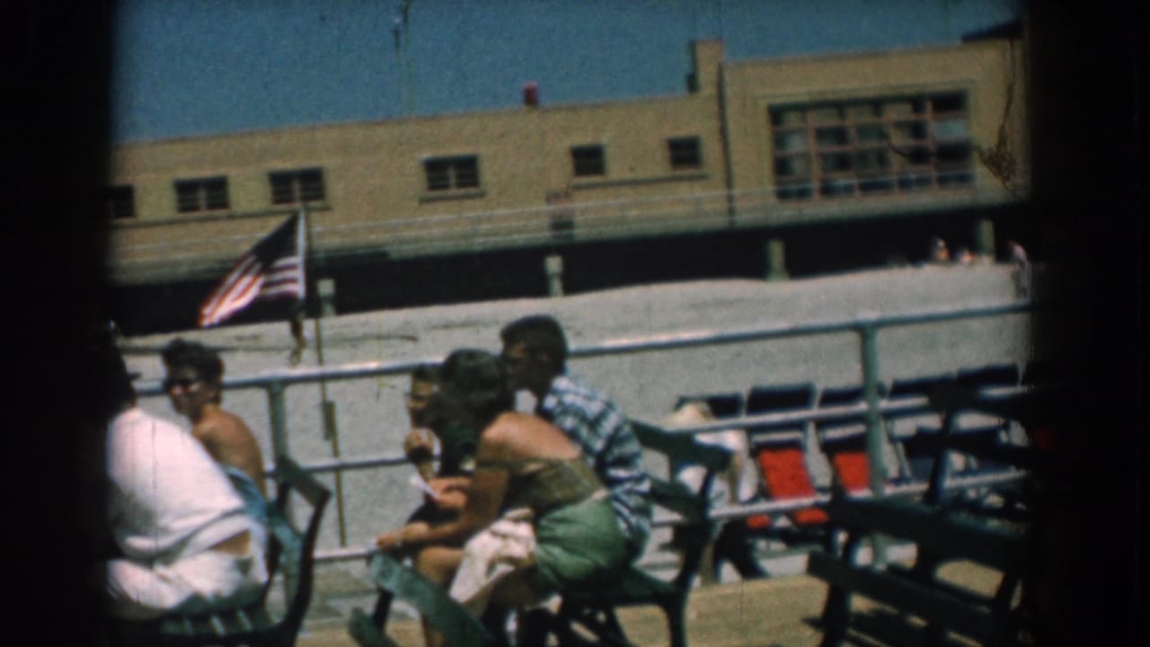 Adults relax on green metal benches at a Yorktown, Virginia boardwalk in 1958, with an American flag and beige building behind them.
