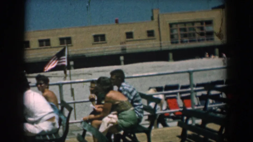 Adults relax on green metal benches at a Yorktown, Virginia boardwalk in 1958, with an American flag and beige building behind them.