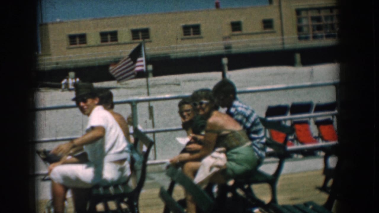 Group of adults on boardwalk benches with flag, railing, and building in background.