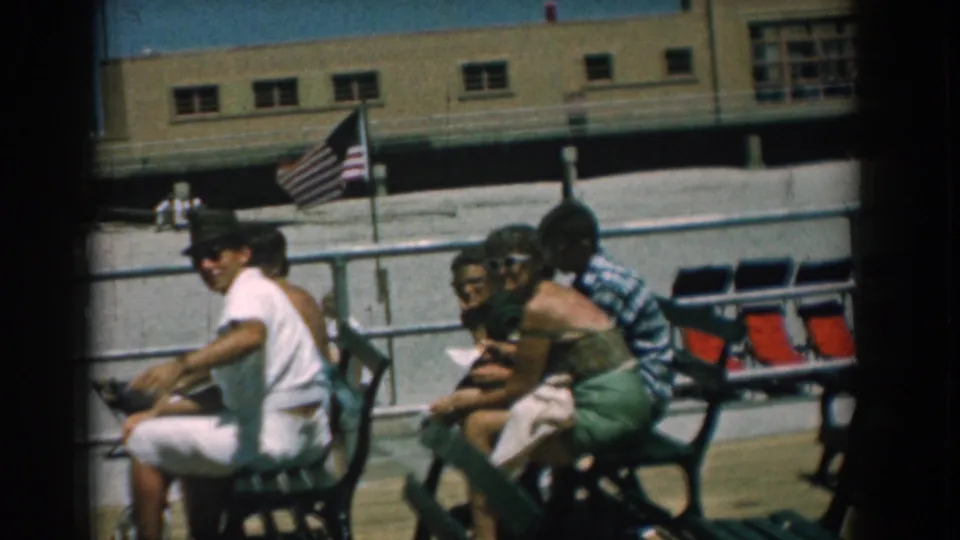 A seated group of adults faces the camera on a sunny Yorktown boardwalk, with an American flag and waterfront building visible behind them.