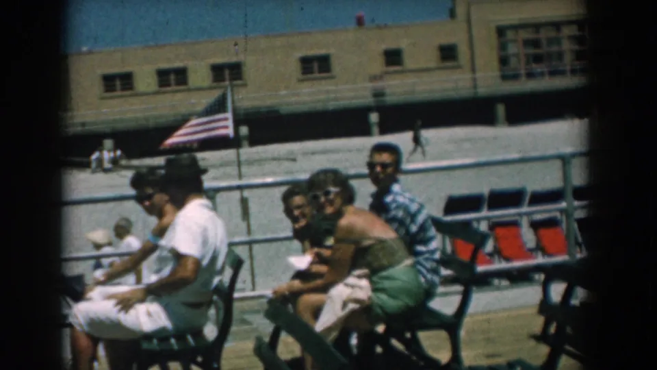 Adults in 1950s summer clothing sit closely on green benches at the Yorktown boardwalk, with the flag and long building in the background.