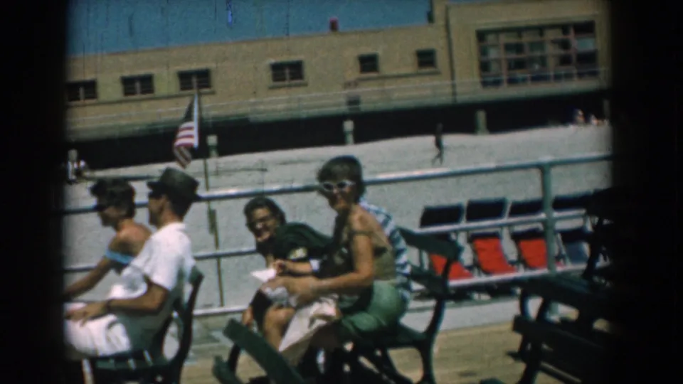 Several adults sit and smile on green benches at the Yorktown boardwalk, with an American flag, railing, red seats, and beige building behind them.