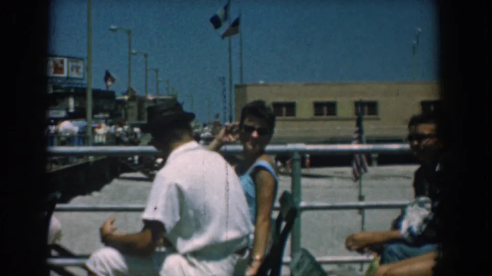 Young adults sit close together near railings and flags in a 1958 Yorktown, Virginia home-movie view of a public boardwalk.