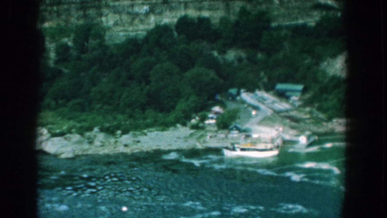 White boat moving near a rocky shoreline below trees and a cliff.