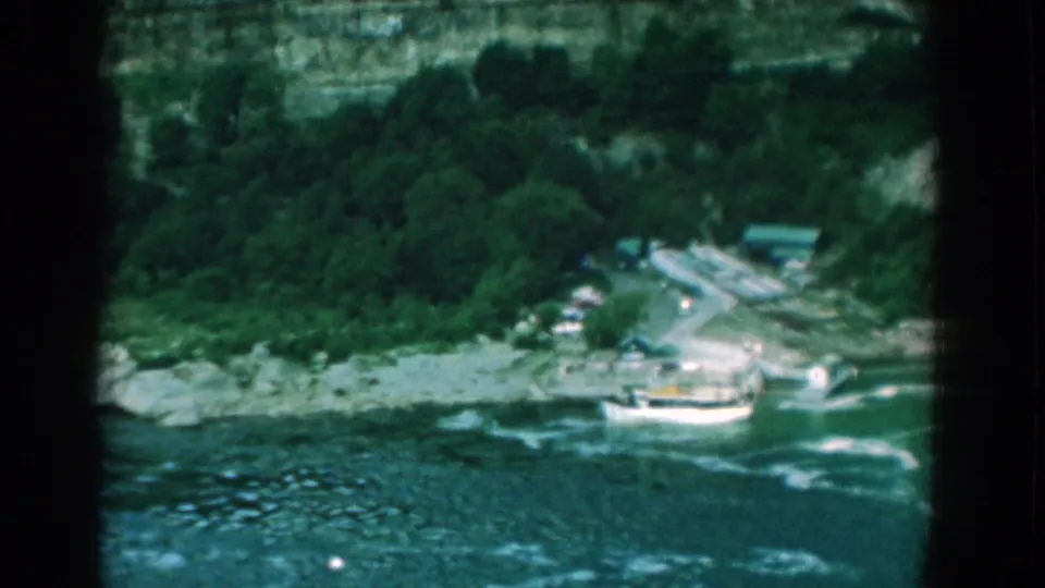 1958 Yorktown, Virginia 8mm frame showing a small white boat on choppy river water near rocks, trees, road, and cliff.