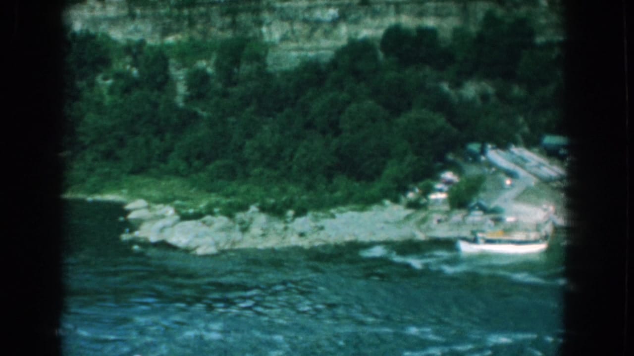 Riverbank with white boat, shoreline rocks, trees, and road at the cliff base.