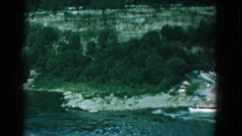 A distant 8mm view of the Yorktown river edge shows rocks, trees, a cliff, roadside cars, and a white boat near the frame edge.