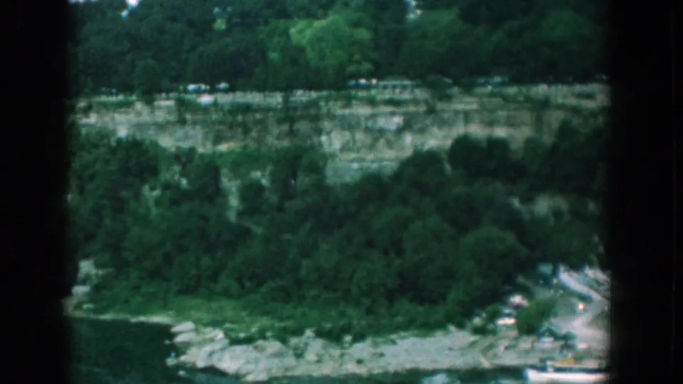 Yorktown river scenery in 1958, with a wooded cliff, pale shoreline rocks, a curving road, parked cars, and a white boat partly visible.