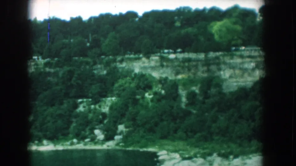 1958 home-movie frame of the Yorktown quarry swimming area, with water, cliff layers, trees, and small distant structures.