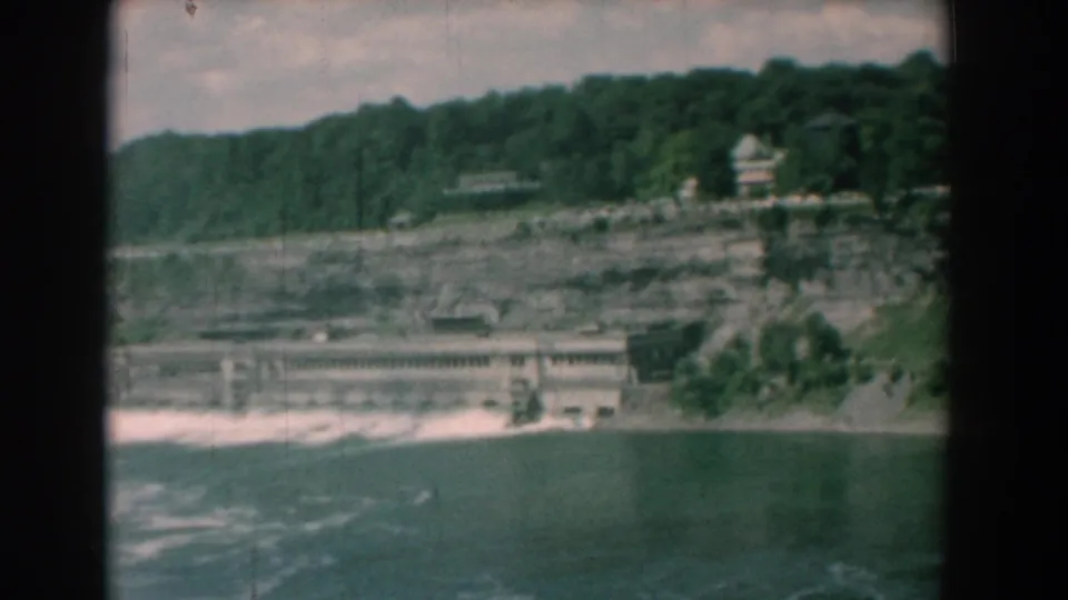 1958 Yorktown 8mm still showing choppy river water, a pale shoreline structure, layered bank, and trees above.
