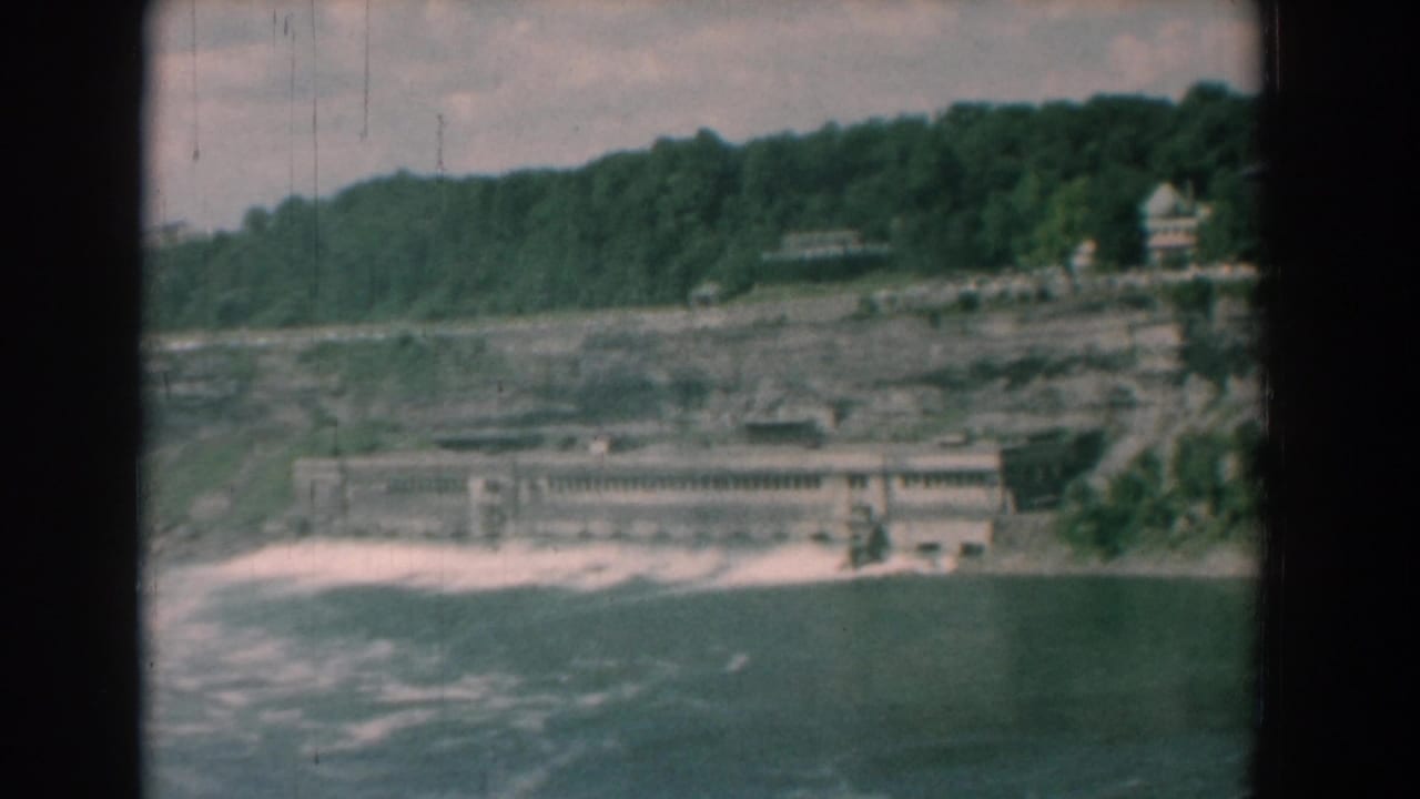 Wide river view with whitewater, concrete structure, rocky bank, and wooded bluff.