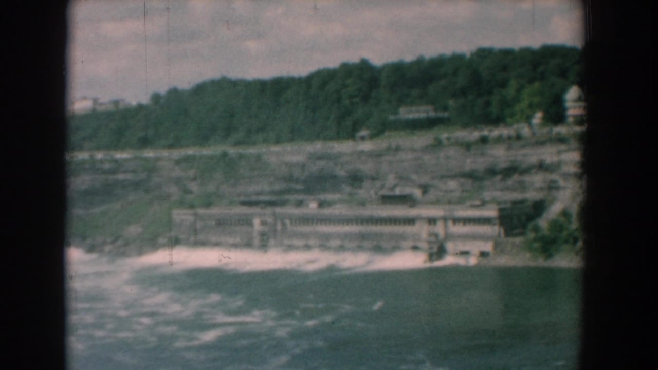 River and low concrete structure seen across water beneath a tree-covered bluff.