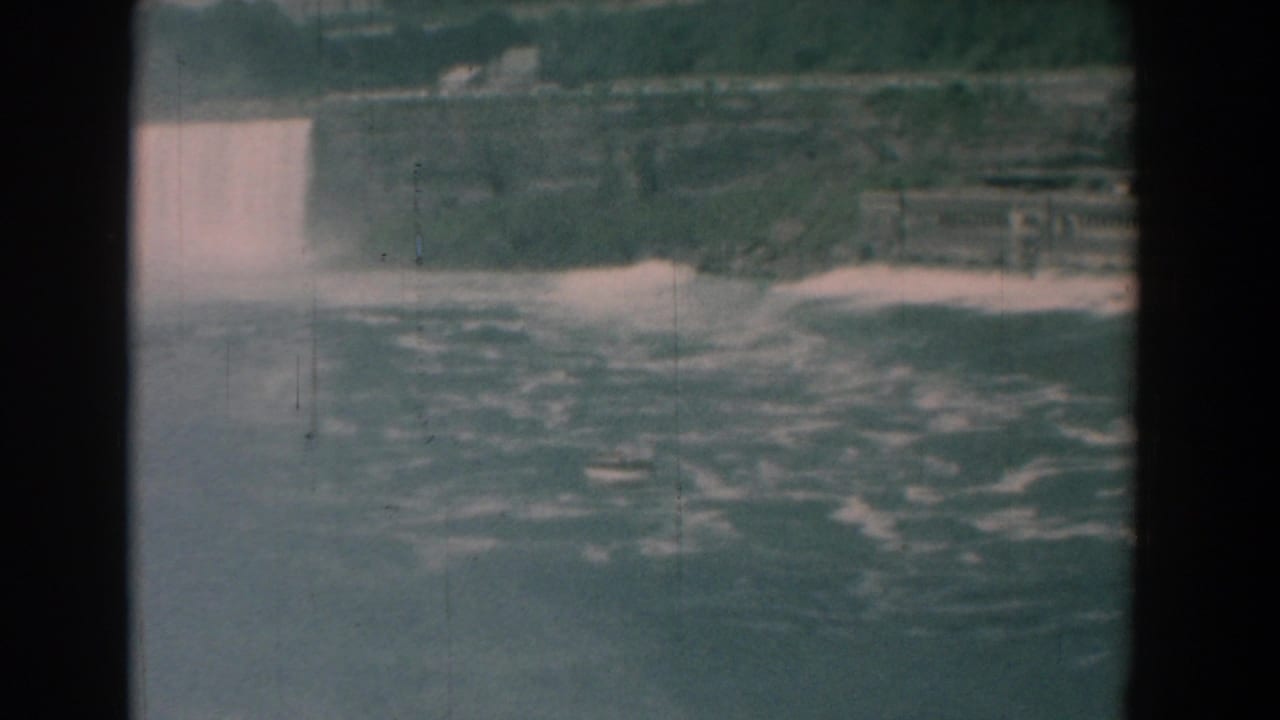 Wide view of water pouring over a drop into a foaming river, with trees and rocks beyond.