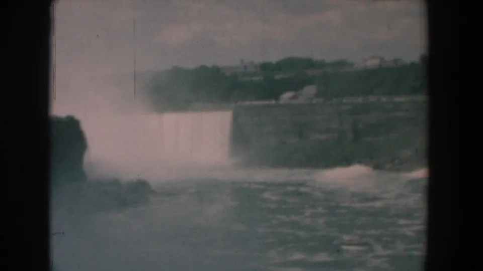 1958 8mm home-movie frame showing a broad waterfall, mist, rocky cliff, and distant shoreline from a visitor viewpoint.