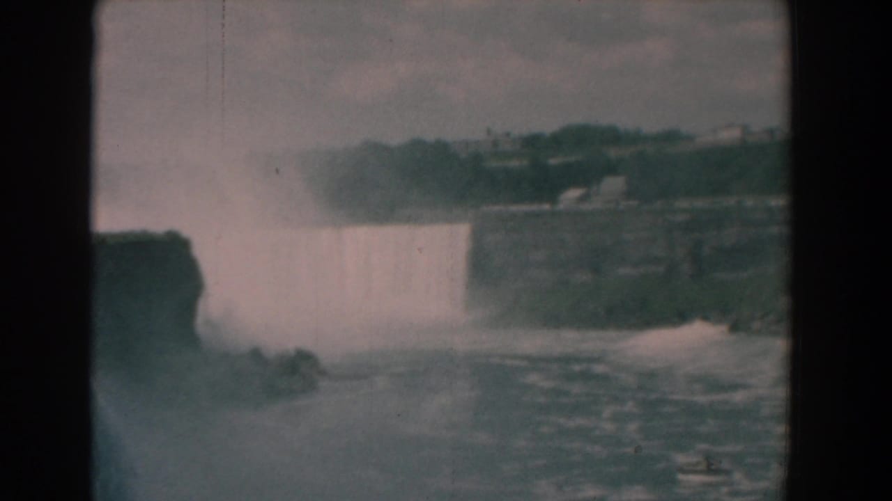 Waterfall and mist seen beyond rough water, framed by dark film edges.
