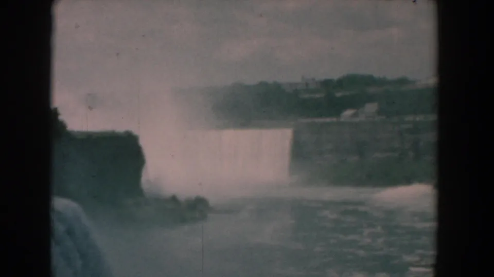 A muted 1958 8mm home-movie frame shows the waterfall from a distance, with spray, dark cliff forms, and rippling water below.