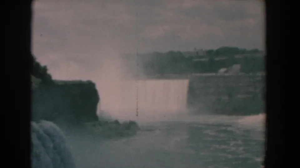 Final 1958 8mm frame showing the waterfall partly obscured by mist, with dark rocks, water texture, and distant greenery visible.