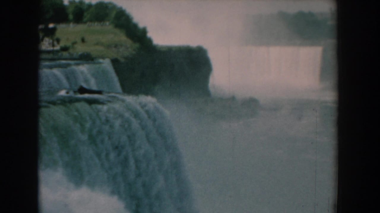 1958 8mm frame of a broad waterfall and rising mist, with a tree-lined grassy bluff in the distance and no visible people.