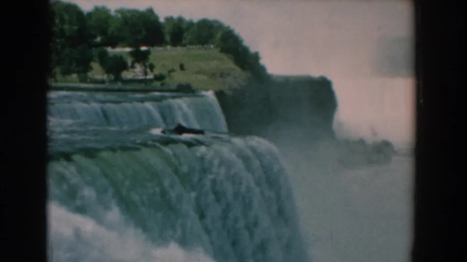 1958 home-movie frame showing rushing water over a broad falls, with mist and a tree-lined bluff beyond.