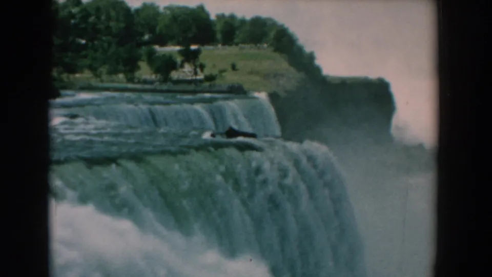 Soft 8mm frame of a wide waterfall pouring into mist, with trees and a grassy overlook visible above the cascade.