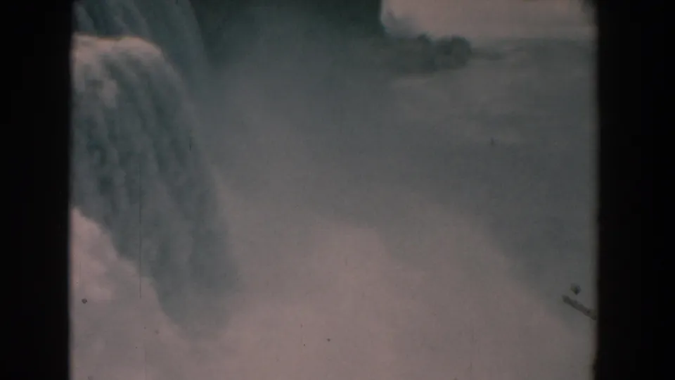 1958 8mm frame of a large waterfall, heavy mist, and distant waterway, filmed from a visitor viewpoint.