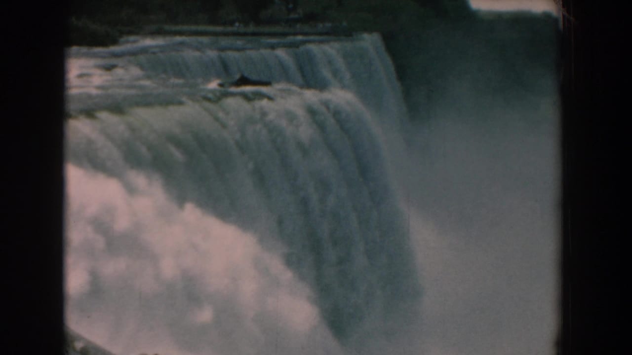 1958 8mm frame of a wide waterfall viewed from above, with heavy water, mist, and a dark tree line beyond.