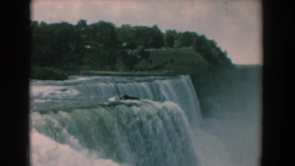 1958 8mm travel frame showing a wide waterfall edge, wooded bank, cloudy sky, and mist rising from below.