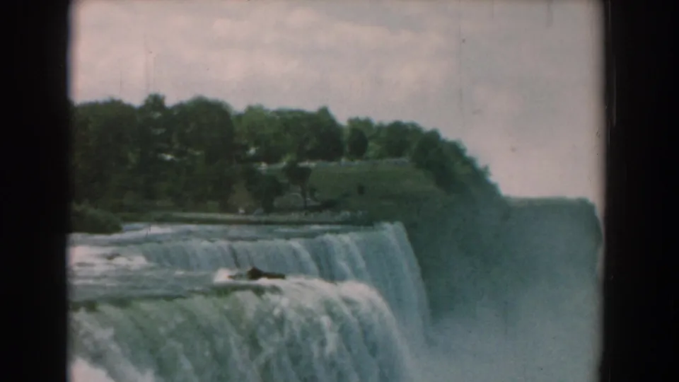 Vintage 8mm frame of the falls from above, showing turbulent water, the cliff rim, trees, and soft mist.