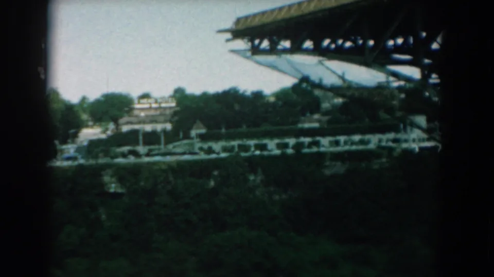 1958 8mm frame shifting from the falls to a distant overlook with a large structure, buildings, trees, and tiny visitors or vehicles.