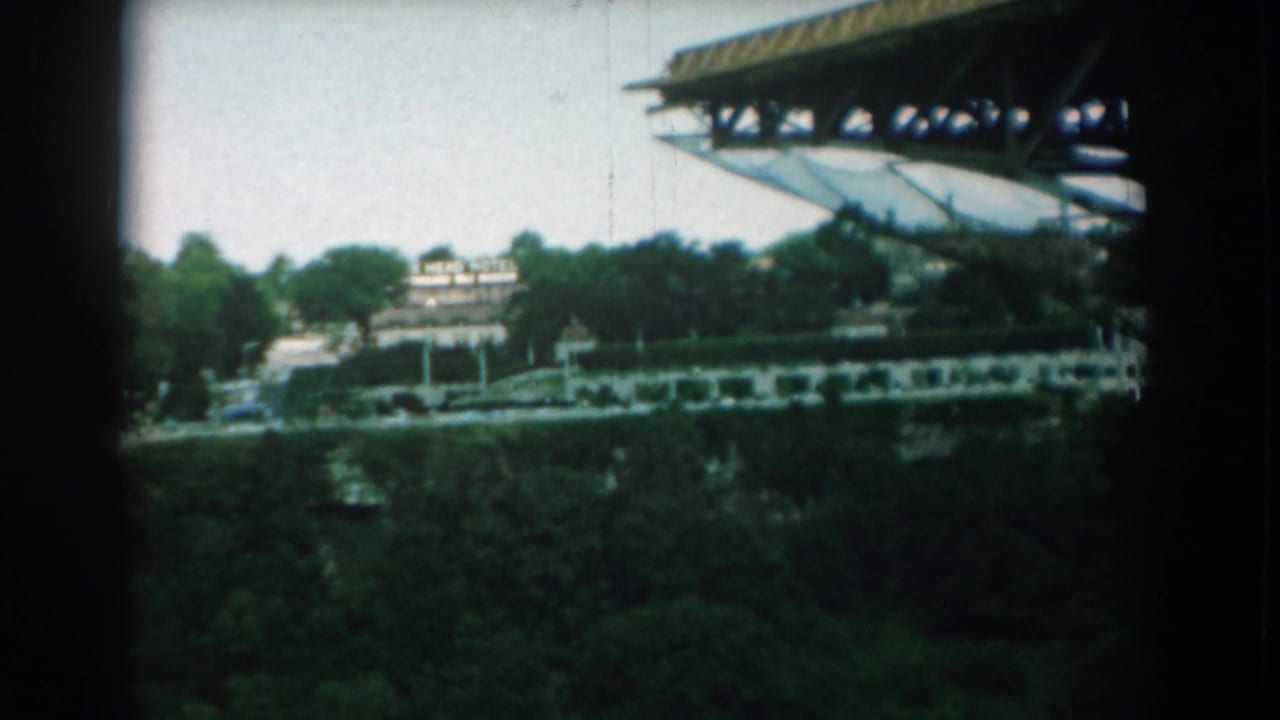 Elevated view past dark frame edges toward trees, road traffic, low buildings, and a large overhead roof structure.