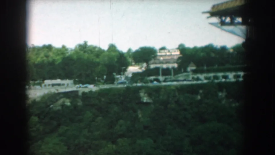 1958 Yorktown visitor footage looking across dense trees and moving traffic toward low roadside buildings under a stadium-like structure.