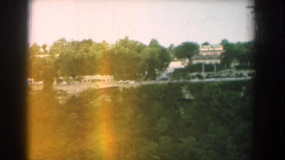 1958 Yorktown home-movie frame showing treetops, a distant built area, pale roadway, and warm exposure flare at the film edge.