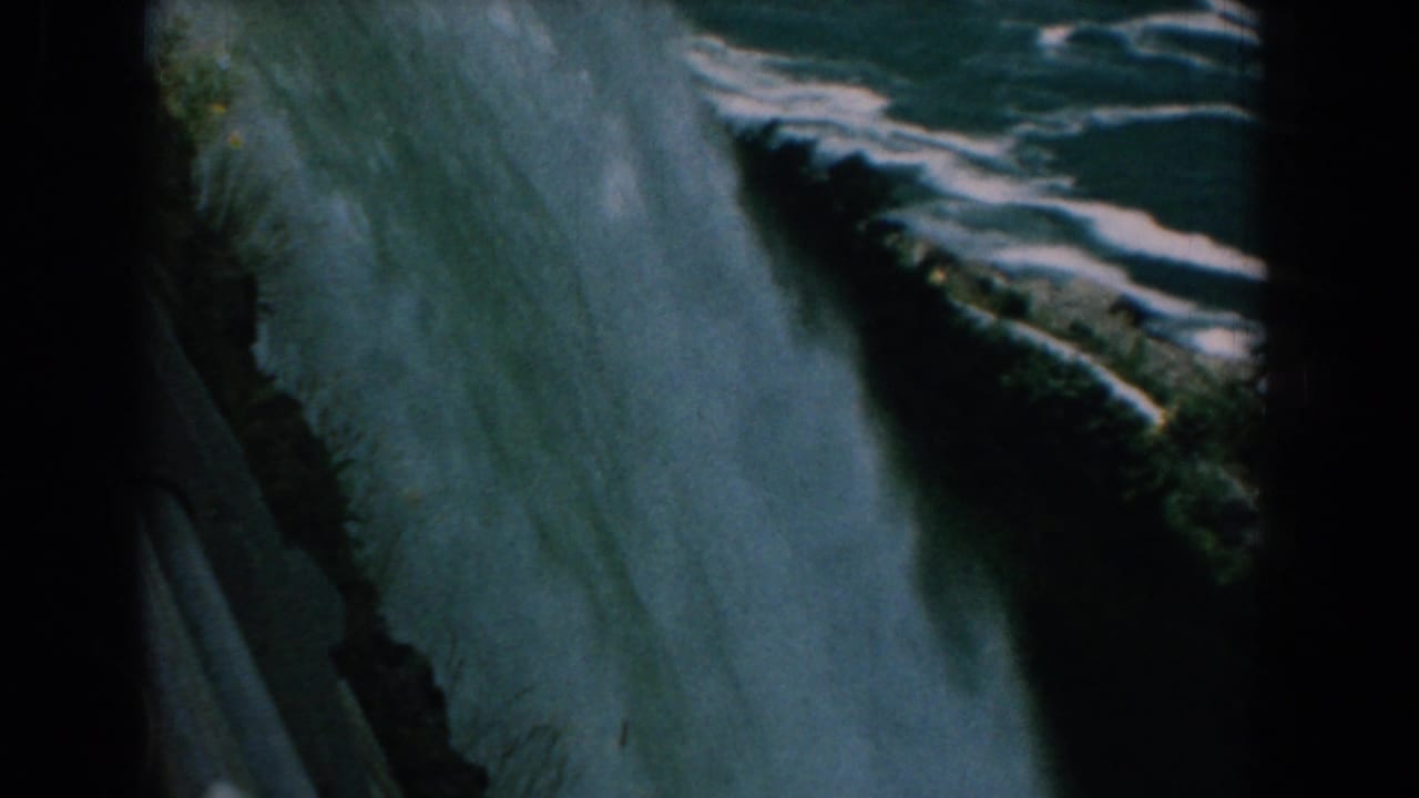Water pours over a steep falls edge beside a rocky green bank.