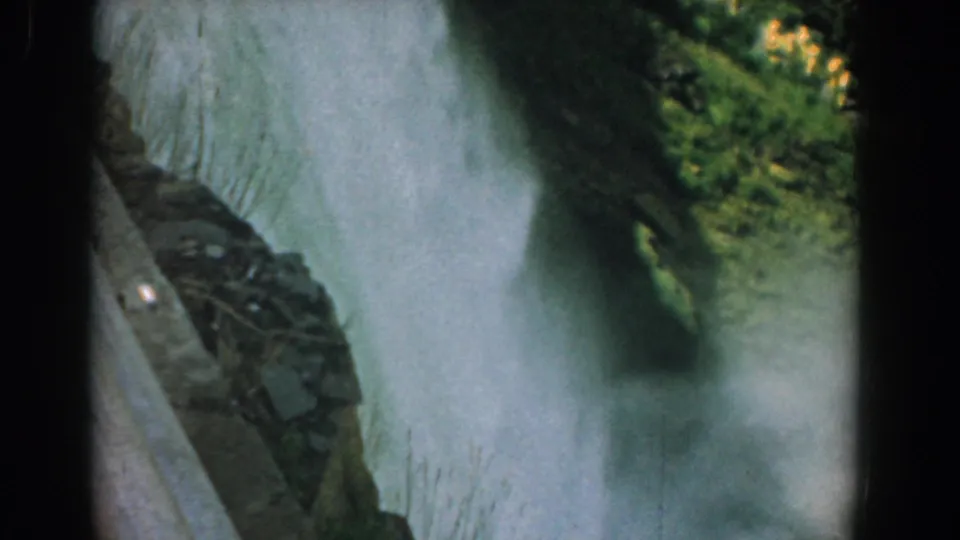 Grainy color 8mm frame showing waterfall spray, a dark rocky outcrop, green vegetation, and a sloped built or stone edge.