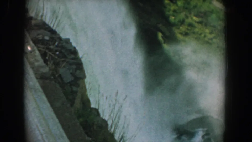 Late-frame 8mm view of the misty waterfall scene, with green vegetation and a dark rock edge softened by motion and spray.