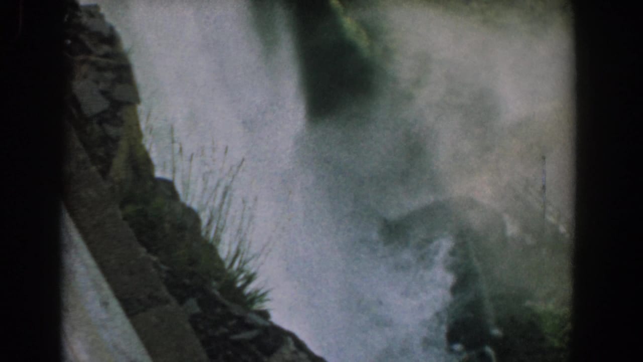 Waterfall and mist framed by rock, greenery, and dark film edges.