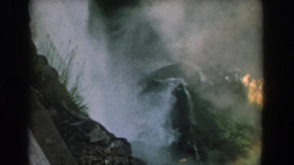 1958 8mm Niagara view with Bridal Veil Falls descending through haze, rock ledges, plants, and indistinct warm shapes near the base.